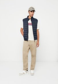 Marine polstret vest med høy krage, brukt over en beige t-skjorte med grafikk, kombinert med beige bukser og hvite sneakers med grå detaljer.