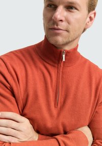 Pull-over orange à demi-fermeture éclair avec un tricot texturé, un col montant et une fermeture éclair argentée. Le tissu semble doux, ajusté avec des manches longues.