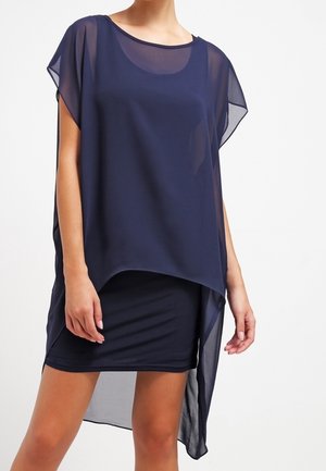 Femme portant une tunique asymétrique transparente bleu foncé sur une robe courte ajustée, debout devant un fond clair uni.