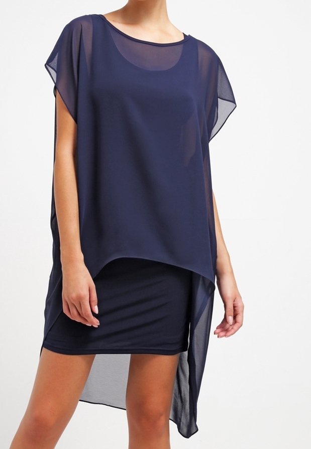 Femme portant une tunique asymétrique transparente bleu foncé sur une robe courte ajustée, debout devant un fond clair uni.