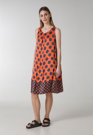 Oranje mouwloze jurk met grote donkerblauwe stippen en een contrasterend donkerblauw patroon aan de onderkant. Gemaakt van een gladde stof, met een ontspannen pasvorm.