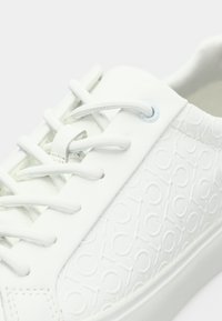 Close-up of white sneaker with textured side panel featuring repeated circular pattern and white laces threaded through metal eyelets.