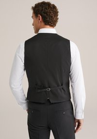 Melnā šķērsjosla ar elegantu dizainu, regulējamu aizmugurējo siksnu un gludu tekstūru, kas valkāta virs baltas krekliņas.