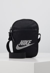 Geantă crossbody din plasă neagră cu închidere cu fermoar, având un logo Nike alb proeminent, curea ajustabilă și suprafață texturată.