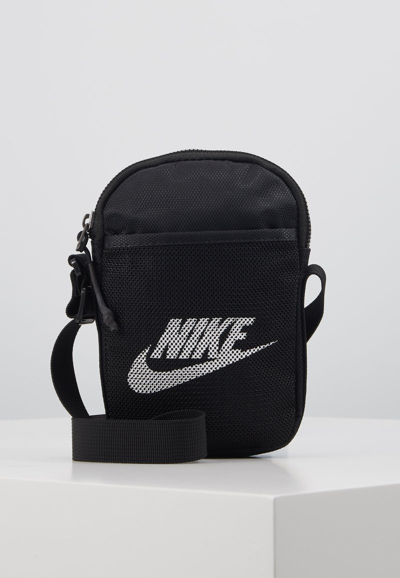 Geantă crossbody din plasă neagră cu închidere cu fermoar, având un logo Nike alb proeminent, curea ajustabilă și suprafață texturată.