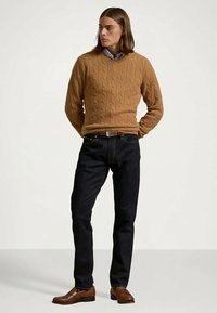 Kabelstrikket genser i en varm tanfarge, kombinert med mørkeblå jeans. Brune skinnsko fullfører antrekket. Minimalistisk design, avslappet stil.