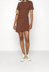 Robe portefeuille marron en tissu extensible, dotée d'un détail de taille noué et de manches courtes. Associée à des baskets montantes beiges.