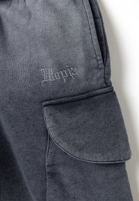 Pantaloni cargo grigi con tessuto testurizzato, dotati di tasca laterale e logo ricamato in una tonalità simile. Cuciture visibili.