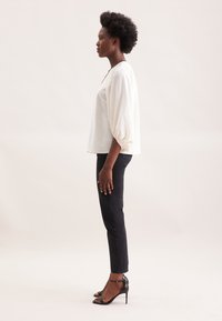Hvid, struktureret bluse med løse, ballonærmer, parret med tætsiddende sorte jeans og strappy sorte højhælede sandaler. Sideprofil.