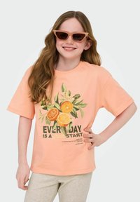 Perzikkleurig t-shirt met korte mouwen heeft een graphic van citrusvruchten en de tekst "IEDERE DAG IS EEN VERS BEGIN." Gecombineerd met lichtgrijze broek.