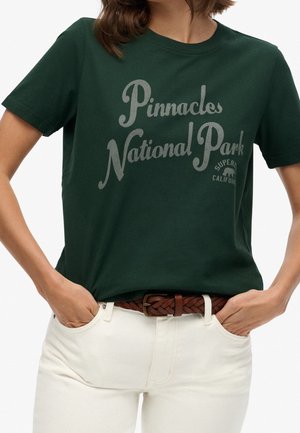 Kobieta ubrana w ciemnozieloną koszulkę z napisem "Pinnacles National Park" i białe spodnie z plecionym brązowym paskiem, ręce w kieszeniach.