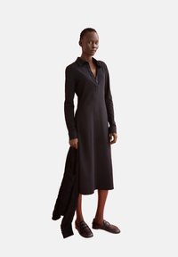 Schwarzes Langarmkleid mit Kragen und drei Knöpfen, kombiniert mit einer gestrickten schwarzen Strickjacke und dunklen Slippern.