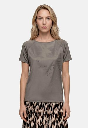 Femme aux cheveux châtain clair portant un t-shirt gris à manches courtes avec un texte subtilement perforé et une jupe à motifs sur un fond uni.
