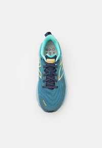 Blå atletisk sko med en texturerad mesh-övre, kontrasterande akvamarina och marinblå snören samt en gul etikett. Har en mönstrad sula för grepp.