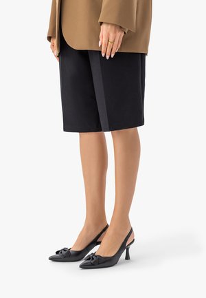 Nogi w czarnych szortach do kolan, brązowej marynarce, czarnych, szpiczastych butach na obcasie z paskiem z tyłu i kokardką, oraz złotych pierścionkach na lewej ręce.