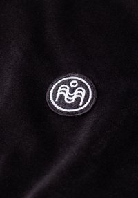 Mustast kangast väikese ümmarguse tikitud plaastriga, millel on valge abstraktse ämbliku kujutis valges ringis.