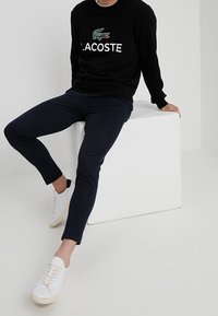 Černá mikina s bílým logem Lacoste, doplněná o námořnické slim-fit kalhoty a bílé tenisky. Hladké textury a minimální designové detaily.