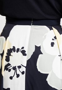 Kukkakuvioitu hame, jossa on suuria mustavalkoisia kukkia vaaleanharmaalla pohjalla, tummansininen vyötärönauha ja laskostettu malli.