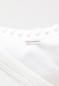 Sneaker bianca in canvas con occhielli metallici e laccetti bianchi. Presenta un piccolo etichetta "Superga" in rosso e blu sul lato. Suola in gomma.