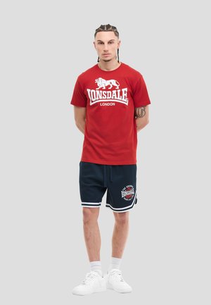 Jonge man met gevlochten haar, gekleed in een rood Lonsdale London T-shirt, donkerblauwe sportshorts met witte rand, witte sokken en witte sneakers.