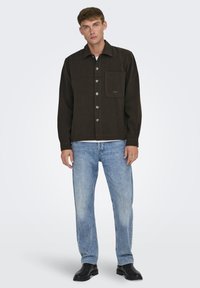 Chaqueta marrón de botones con un bolsillo en el pecho, con botones plateados y una tela texturizada, combinada con jeans azul claro y zapatos negros.