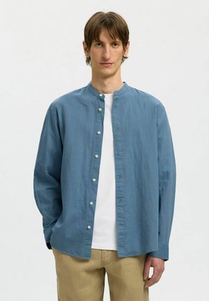 Młody mężczyzna o brązowych włosach ma na sobie niebieską koszulę na guziki, pod którą znajduje się biała koszulka, oraz beżowe spodnie, stoi na jasnym, jednolitym tle.