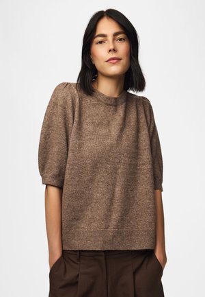 Femme aux cheveux noirs mi-longs portant un pull marron à manches courtes et un pantalon marron foncé, les mains dans les poches, sur fond blanc.