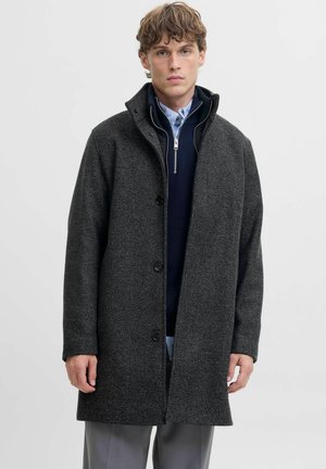 Manteau en laine gris mélangé avec une finition texturée, un col haut et cinq boutons noirs. Porté sur un pull à zip bleu et une chemise à col.