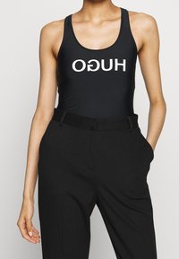 Traje de baño negro con el atrevido texto blanco "HUGO" en el frente, fabricado con un material suave, combinado con pantalones de tiro alto negros y ajustados.