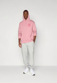Sweatshirt rosa com capuz, bolso frontal e logótipo, combinada com calças de fato de treino cargo cinza claro. Modelo usa sapatos desportivos brancos, em pé sobre um fundo simples.