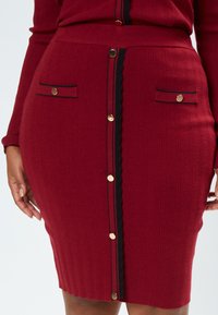 Roter gerippter Bleistiftrock mit schwarzem Saum, ausgestattet mit einer vorderen Knopfverschlüsse und zwei vorderen Taschen mit goldenen Akzenten. Texturiertes Strickmaterial.