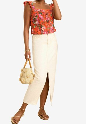 Femme portant un haut sans manches orange à motifs floraux, une jupe taille haute crème avec une fente à l'avant, tenant un sac à main en paille texturée, et portant des sandales dorées.