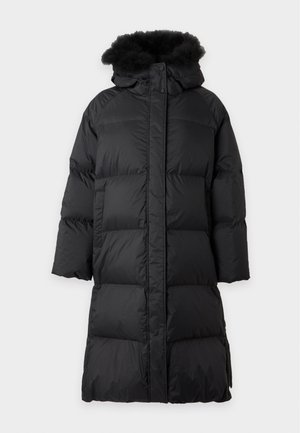 Femme portant un long manteau noir matelassé avec capuche en fourrure, un pull rouge à col en V, un jean bleu avec des fentes à l'avant, et des talons noirs à bout pointu, debout sur un fond blanc.