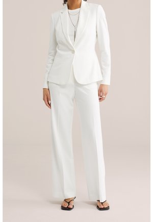 Mujer con traje blanco a medida con blazer de un solo botón, pantalones de pierna ancha, collares plateados en capas y sandalias negras con punta abierta.