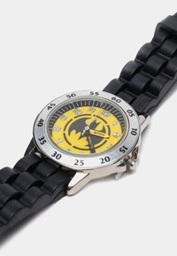 Montre jaune avec un logo de chauve-souris noir sur le cadran. Lunette en argent avec des marqueurs de minutes. Bracelet en silicone noir texturé, forme ronde.