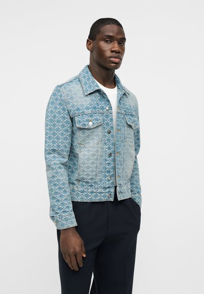 Νεαρός άνδρας με σκούρο δέρμα φοράει ένα ανοιχτό γαλάζιο τζιν μπουφάν με σχέδια πάνω από ένα άσπρο πουκάμισο και σκούρο παντελόνι, στέκεται μπροστά σε ένα απλό φόντο.