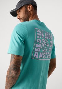 Mand med teal T-shirt med stor, stiliseret firkantet tekst, der står "Scotch & Soda 1985 Amsterdam" på ryggen, og en mørk kasket.