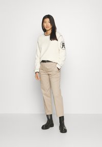 Maglione bianco a trama con accenti neri, pantaloni beige e stivali neri alla caviglia. Il maglione presenta un logo sulla manica sinistra.