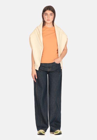 Beige oversized sweatervest over en orange t-shirt, parret med vide ben mørke denim jeans og multicolore sneakers. Simpelt design, bløde teksturer.