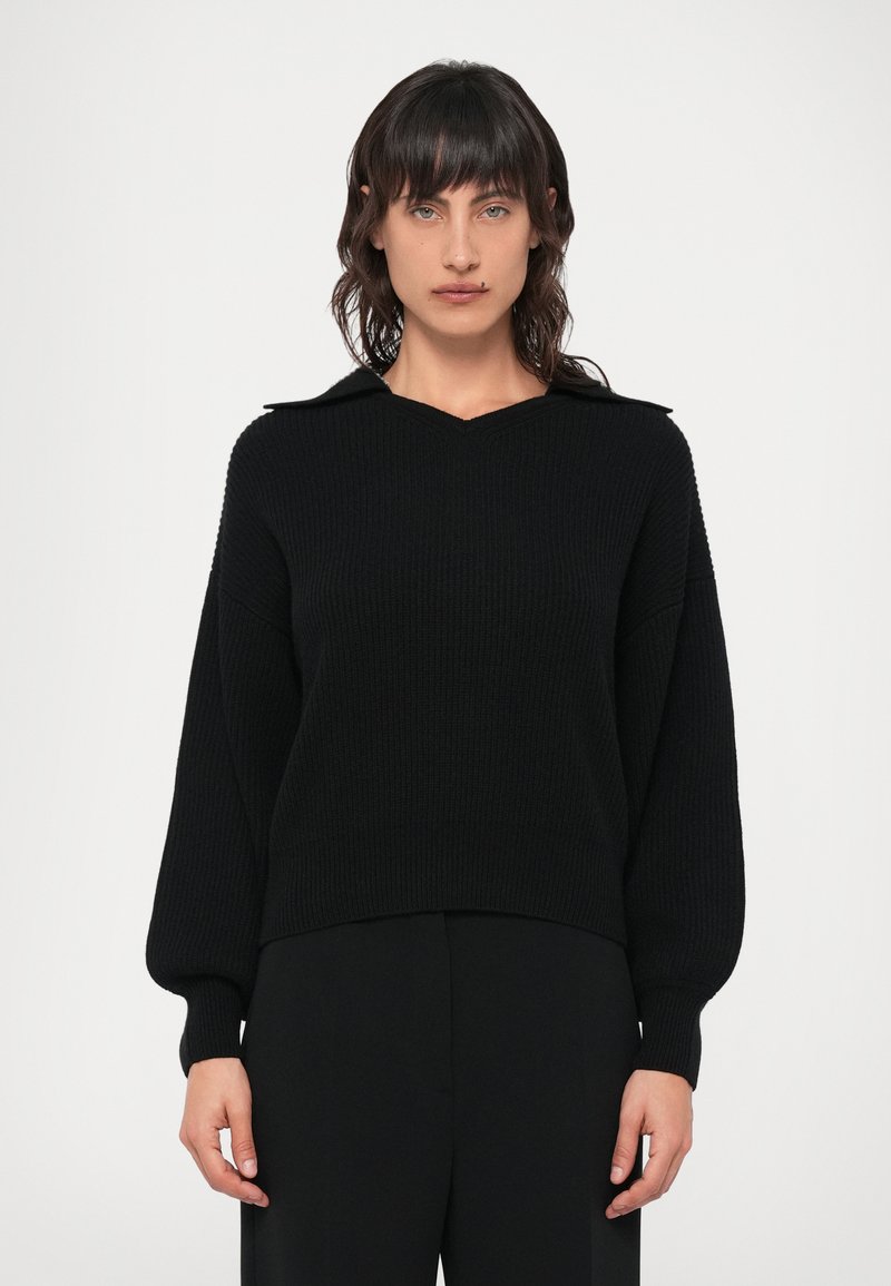 Fekete kötött pulóver V-nyakkal és galléros kialakítással. Laza ujjak és bordázott textúra jellemzi, testre szabott fekete nadrággal párosítva.