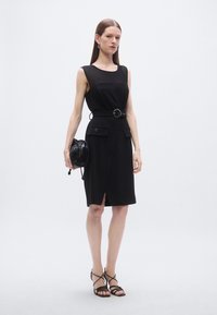 Svart ermeløs kjole med belte i midjen, sidelommer og splitt foran. Kombinert med en svart snorveske og sandaletter med stropper.