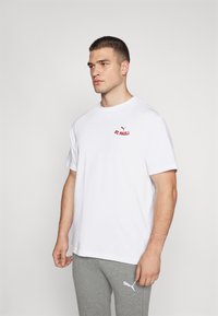 Balta īsām piedurknēm t-kreklā ar apaļu sarkanu "St. Pauli" logotipu uz krūtīm, apvienots ar pelēkiem biksēm, uz vienkārša fona.