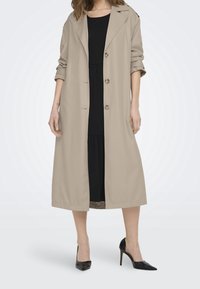 Krémový trench coat s pročesávacím límcem, zapínáním na knoflíky a širokými rukávy, kombinovaný s černými šaty a černými vysokými podpatky.