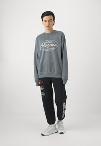 Grå hettegenser med hvit logo tekst, kombinert med svarte joggebukser med fargerike lapper og hvite tekstdetaljer, fullført med hvite sneakers.