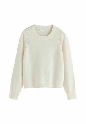 Vrouw met kort zwart haar die een witte trui, losse zwarte krijtstreepbroeken en zwarte sandalen draagt, lopend met handen in de zakken.