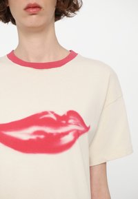 Cream colored t-shirt featuring a large bright pink lip print on the front and a contrasting pink ribbed neckline. Soft fabric texture.