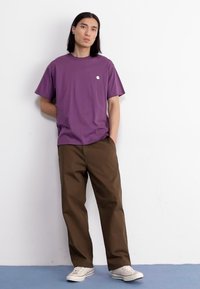 Paarse katoenen t-shirt met korte mouwen en een klein logo op de borst, gecombineerd met bruine losse broek en witte sneakers.