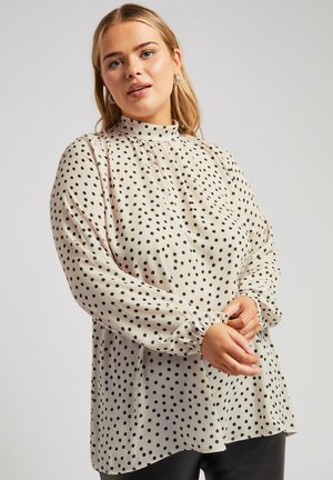 Blusa beige chiaro con pois neri, colletto alto, maniche lunghe con polsini arricciati e una vestibilità ampia, realizzata in un tessuto liscio e fluente.