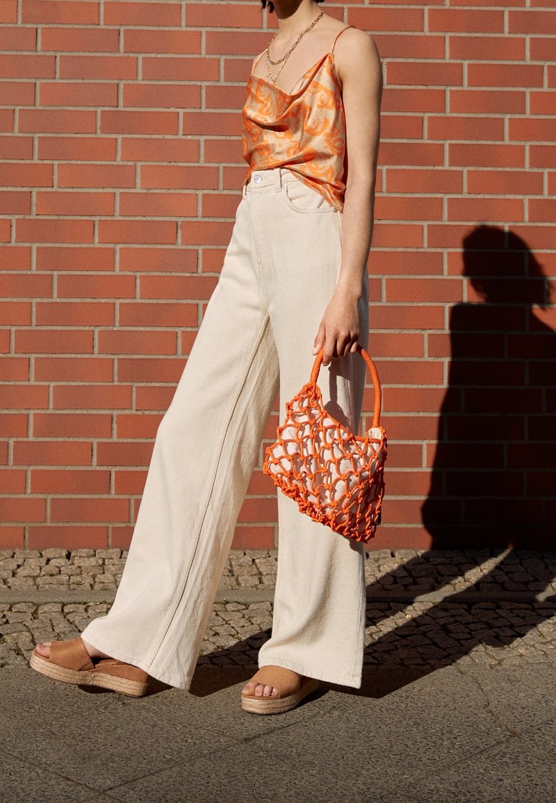 Femme portant un haut en satin orange et un pantalon blanc à jambes larges, tenant un sac en filet orange, debout contre un mur en briques rouges.