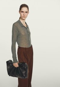 Graues Langarm-Hemd mit Knöpfen und Kragen, kombiniert mit braunen, plissierten Hosen. Hält eine große, schwarze Ledertasche mit verwaschener Textur.